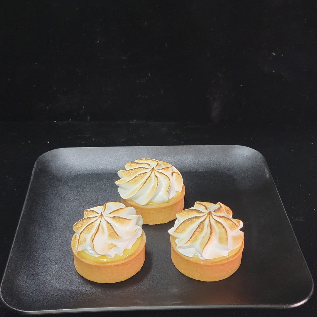 "Three gourmet lemon meringue tarts with toasted, swirled meringue peaks on a matte black square serving plate, shot against a solid black studio background."