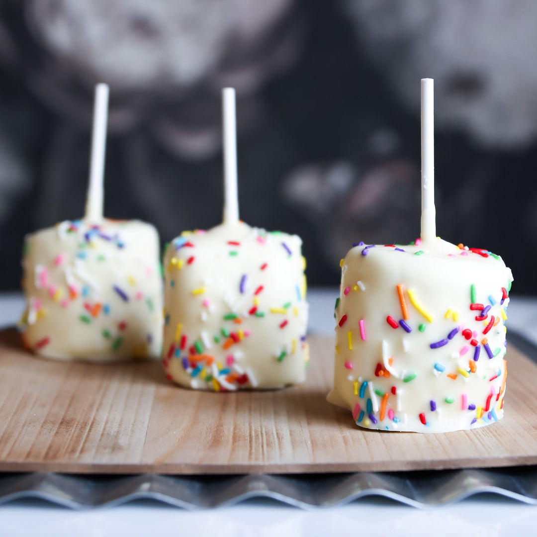 Three marshmallows on sticks with colorful sprinkles on a wooden board.