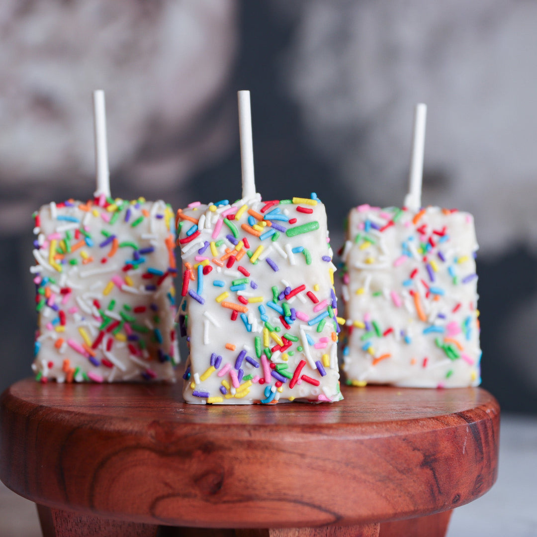 Three marshmallow pops with colorful sprinkles on a wooden surface.