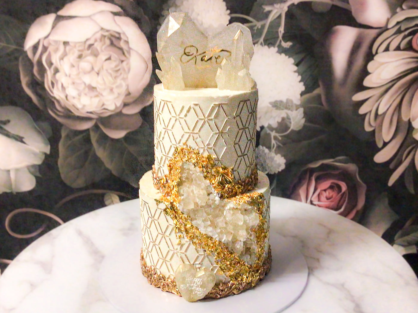 geode sugar crystals delicately covering a carved out piece to create a geode crystal illusion. Geodes are then outlined with 24 Carat gold leaf from India. Another later of sophistication is added to the cake with hand painted gold hexagon patterns around the cake. Last, this cake is embellished with isomalt sugar crystals and hearts, gold painted fondant scroll borders.