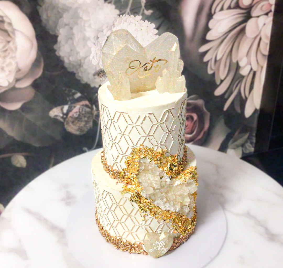 geode sugar crystals delicately covering a carved out piece to create a geode crystal illusion. Geodes are then outlined with 24 Carat gold leaf from India. Another later of sophistication is added to the cake with hand painted gold hexagon patterns around the cake. Last, this cake is embellished with isomalt sugar crystals and hearts, gold painted fondant scroll borders.