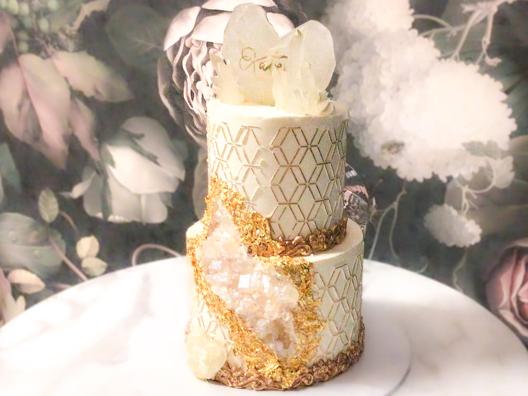 geode sugar crystals delicately covering a carved out piece to create a geode crystal illusion. Geodes are then outlined with 24 Carat gold leaf from India. Another later of sophistication is added to the cake with hand painted gold hexagon patterns around the cake. Last, this cake is embellished with isomalt sugar crystals and hearts, gold painted fondant scroll borders.